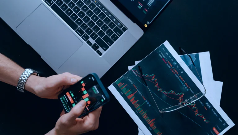 Vue rapprochée d'un investisseur analysant des graphiques financiers sur un smartphone et un ordinateur portable, avec des documents imprimés et des lunettes posées sur la table.